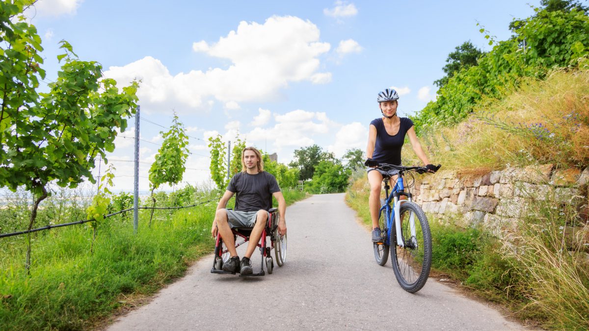 Young couple enjoying time outdoors. She is cycling, he is sitting in a wheelchair.