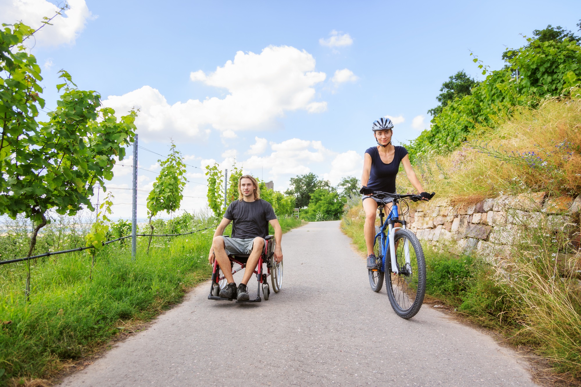 Young couple enjoying time outdoors. She is cycling, he is sitting in a wheelchair.